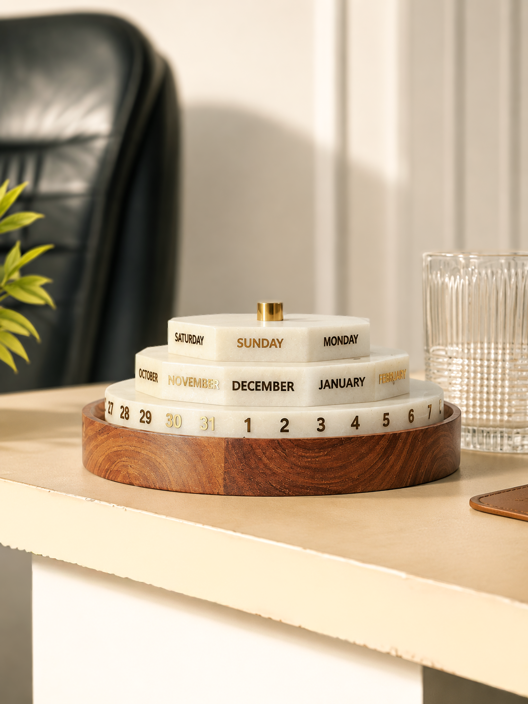 A tiered monthly calendar display on a wooden table, featuring cream-colored stone-like rings labeled with days of the week and months, with a gold-colored top knob. The background shows a blurred black leather chair and a glass on a beige table.