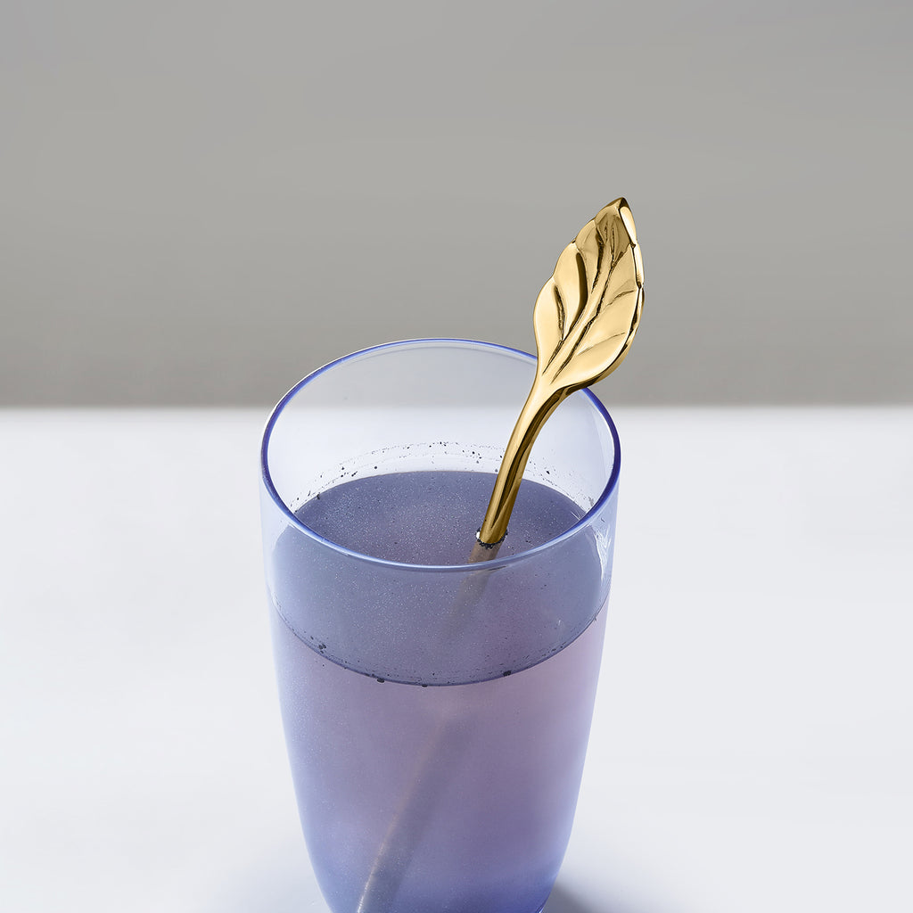 Gold leaf spoon in a glass of purple liquid on a light gray background