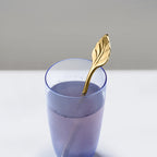 Gold leaf spoon in a glass of purple liquid on a light gray background