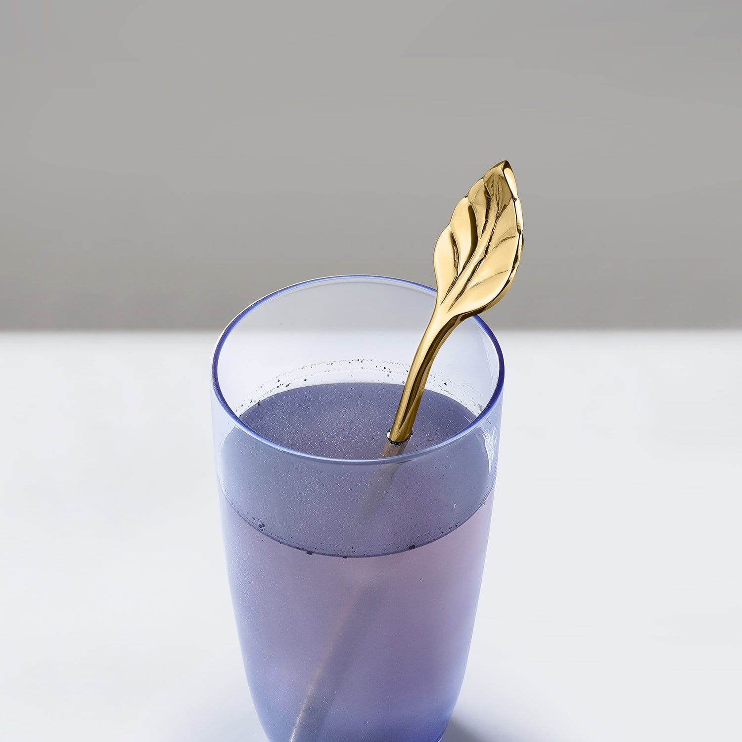 Gold leaf spoon in a glass of purple liquid on a light gray background