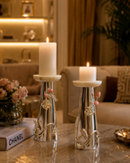 Two silver metal candlestick holders with white marble tops and small pink floral accents, each holding a lit white pillar candle, displayed on a polished marble table in a warm, upscale living room setting.