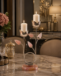 A decorative metal candle holder with pink rose-shaped accents and a pink stone base, holding two lit white candles on a marble dining table in an elegant interior setting.