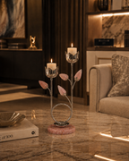 A decorative metal candle holder with pink rose-shaped petals and a pink marble base, holding two lit white candles, placed on a polished marble table in a modern living room setting.