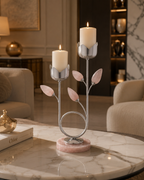 A decorative metal candle holder with leaf-shaped pink stone accents and a round pink marble base, shown on a marble table in a softly lit living room. Two white taper candles are lit in the holder.